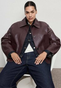 Bordowa kurtka z eko skóry z kołnierzem z wycięciem, czarnymi guzikami i bocznymi kieszeniami, noszona na koszuli denimowej i ciemnych dżinsach.