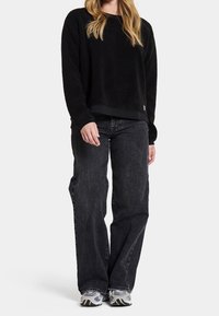 Donna che indossa un maglione in pile nero, jeans neri larghi e sneaker grigie, in piedi con le mani rilassate contro uno sfondo chiaro e semplice.