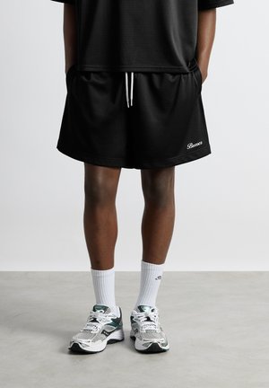 Person iført sorte atletikshorts med hvid snøre og hvide sokker, kombineret med hvide og grønne løbesko, stående på gråt gulv.