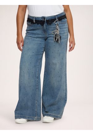 Weit geschnittene blaue Jeans mit kleinen schwarzen Punkten, einem schwarzen Spitzenbund und einem Schlüsselanhänger, der an der Gürtelschlaufe hängt.