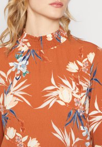 Blusa color ruggine con motivo floreale in crema, blu e verde. Presenta un colletto alto con volant e un tessuto morbido e testurizzato.