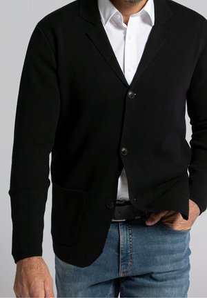 Mann trägt schwarzen, geknöpften Cardigan über weißem Hemd und blauer Jeans, mit einer Hand in der Tasche, vor einem schlichten grauen Hintergrund.