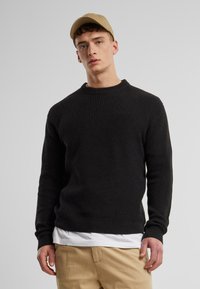 Young man wearing a beige cap, black ribbed sweater over a white shirt, and beige pants against a plain light gray background.