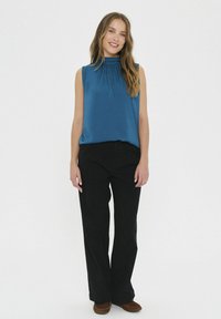 Top blu senza maniche con scollatura arricciata, abbinato a pantaloni neri larghi. Scarpe slip-on marrone scuro completano l'outfit.