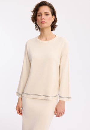 Kobieta w kremowej bluzce z długim rękawem i dopasowanej spódnicy, z subtelnym czarnym przeszyciem na rękawach i dole, stoi na białym tle.