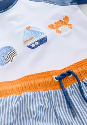 Baddräkt med en vit överdel med blå detaljer och broderade motiv av en val, segelbåt och krabba. Ränder med blå och vita elastiska shorts med orange midja.