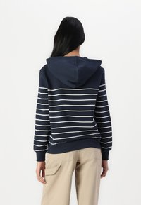 Granatowa bluza z kapturem z białymi poziomymi paskami, wykonana z bawełny, z regulowanym kapturem, z prążkowanymi mankietami i dołem; noszona z beżowymi spodniami cargo.