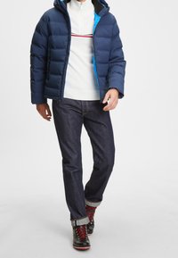 Námořnická péřová bunda s modrým podšívkou, nošená přes bílý svetr s červenými pruhy, doplněná tmavými džínami a černými šněrovacími botami.