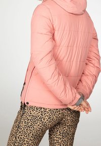 Personne portant une doudoune rose clair avec une capuche et un pantalon à imprimé léopard, debout les mains jointes derrière le dos.