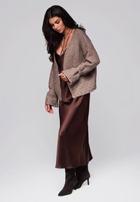 Brauner, strukturiertes Cardigan mit Glockenärmeln über einem satinbraunen Slipkleid und schwarzen High-Heels. Eine einfache und elegante Outfit-Kombination.