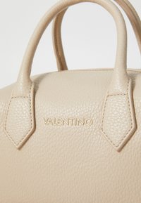 Borsa a mano in pelle beige con superficie texturizzata, doppi manici e logo "Valentino" in rilievo dorato. Presenta dettagli geometrici sui manici.