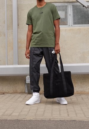 Osoba nosząca oliwkowo-zielony t-shirt, czarne spodnie z logo Nike, białe sneakersy, trzymająca dużą czarną torbę na tle betonowej ściany.