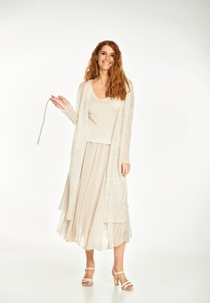 Uśmiechnięta kobieta o długich rudych włosach, ubrana w beżowy długi kardigan, dopasowany top, zwiewną spódnicę oraz buty na obcasie z odkrytymi palcami, stoi na białym tle.