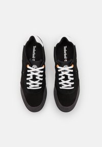 Baskets en daim noir avec des lacets et des accents de talon blancs, dotées d'une semelle en caoutchouc grise. Logo Timberland sur la languette et sur le côté.