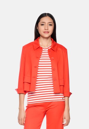 Frau trägt leuchtend rote Jacke über einem weiß-rot gestreiften Hemd mit passenden roten Hosen, steht vor einem einfarbigen hellen Hintergrund.