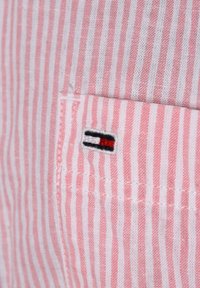 Randig tyg i rosa och vitt, med en liten broderad logotyp i svart, vitt och rött. Närbild på sömnadsdetalj.