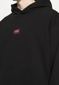 Sweat à capuche noir en coton, avec un petit patch logo rouge sur la poitrine et une poche kangourou sur le devant. Texture douce.