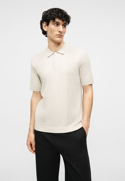 Νέος άνδρας με σγουρά σκούρα μαλλιά φοράει μπεζ κοντομάνικο πόλο πουκάμισο σε συνδυασμό με μαύρο παντελόνι, στέκεται μπροστά σε απλό ανοιχτό φόντο.