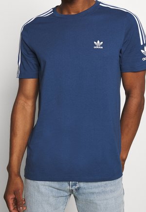 Mężczyzna noszący granatową koszulkę Adidas z białymi paskami na ramionach i rękawach, w połączeniu z jasnoniebieskimi dżinsami.