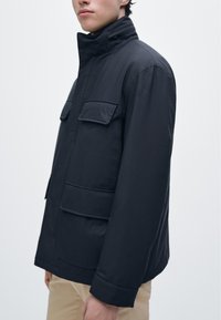 Zwart gewatteerd jack met hoge kraag, twee borstzakken en een gladde textuur. Heeft een aansluitend silhouet en knoopmanschetten.