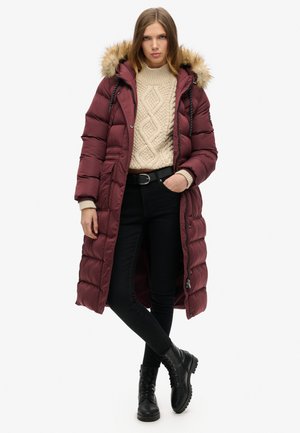 Bordeaux lange pufferjas met een met bont afgewerkt capuchon, beige trui met zigzagpatroon, zwarte broek en zwarte veterschoenen. Bevat een ritssluiting en zakken.