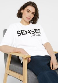 White cotton t-shirt with bold black text "SENSES .THE LABEL" displayed prominently; model seated in a light wood and gray upholstered chair.