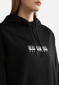 Sudadera negra con capucha que presenta un logo impreso en blanco en el pecho, con cordones en la capucha y un tejido suave al tacto. Diseño sencillo con corte relajado.