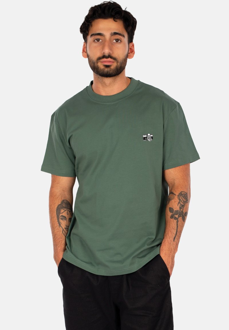 T-shirt in cotone verde con collo tondo e maniche corte. Presenta una piccola grafica nera di una macchina fotografica sul lato sinistro del petto.