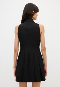 Černé bezramínkové šaty s vysokým límcem, přiléhavým živůtkem a rozevlátou plisovanou sukní. Hladká látka s elegantním designem a minimálním zdobením.