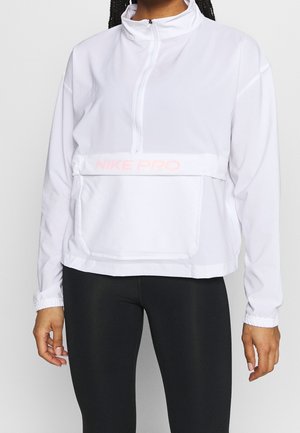 Personne portant un haut blanc Nike Pro à manches longues avec fermeture éclair à mi-hauteur et des leggings noirs, debout les bras détendus le long du corps.