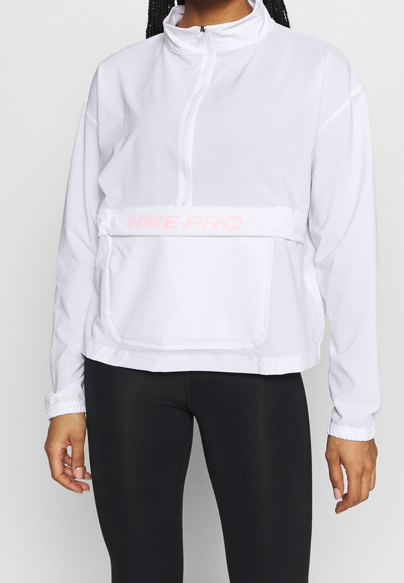 Personne portant un haut blanc Nike Pro à manches longues avec fermeture éclair à mi-hauteur et des leggings noirs, debout les bras détendus le long du corps.
