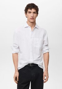 Weißes Langarmhemd aus Leinenstoff mit einem Button-Down-Kragen und einer einzelnen Tasche; kombiniert mit schwarzen Hosen und einem schwarzen Gürtel.