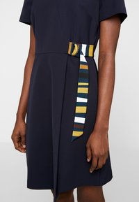 Femme portant une robe bleu marine à manches courtes et une ceinture en tissu rayée multicolore maintenue par une boucle ronde à la taille.