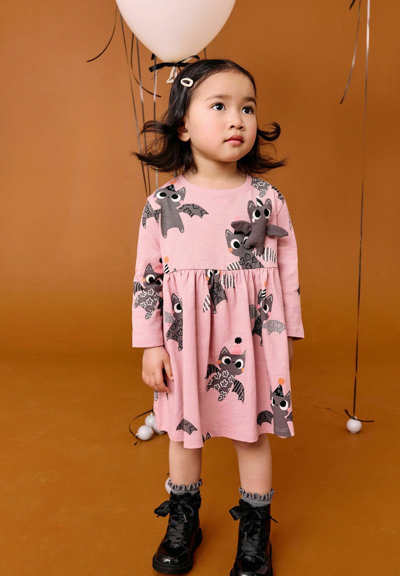 Junges Mädchen mit dunklem Haar steht, trägt ein pinkes Kleid mit Fledermausmuster und schwarze Stiefel und hält einen weißen Ballon vor braunem Hintergrund.