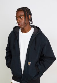 Young man with braids wearing a black hooded jacket over a white shirt, looking to the left with hands in jacket pockets.