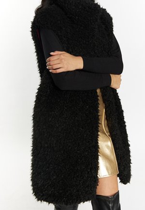 Person trägt eine lange schwarze, flauschige Weste, schwarze Langarmshirts, einen metallisch goldenen Rock und schwarze kniehohe Stiefel mit verschränkten Armen.