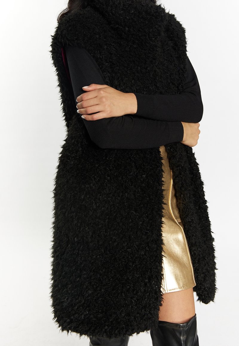 Personne portant un long gilet noir duveteux, des manches longues noires, une jupe métallique dorée et des bottes noires montantes, bras croisés.