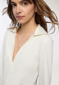 Donna con capelli castano chiaro che indossa una blusa bianca con colletto a V profonda su uno sfondo chiaro.