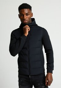 Crna podstavljena zip dukserica s visokim ovratnikom, ravnim šavovima i podesivom kapuljačom na vezanje. Ima glatku vanjštinu i rebraste manžete.