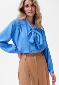 Jasnoniebieska bluzka z kokardą przy kołnierzyku, z długimi rękawami i marszczonymi detalami. Połączona z brązowymi spodniami o wysokim stanie z guzicznymi dodatkami.