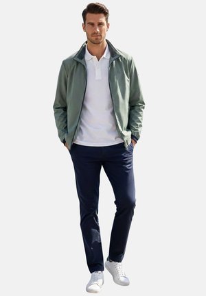 Hombre de pie con las manos en los bolsillos, vistiendo una chaqueta verde con cremallera, camisa polo blanca, pantalones azul marino y zapatillas blancas sobre fondo blanco.