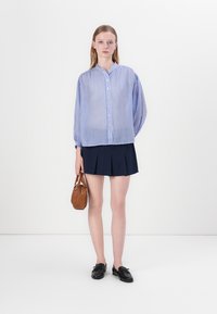 Sininen ja valkoinen raidallinen pusero, napitettu etuosa, yhdistetty tummansinihin laskostettuun hameeseen. Pienessä ruskeassa laukunpidkkeessä, mustat kengät täydentävät asua.