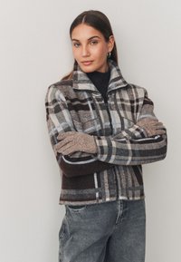 Veste zippée à carreaux marron et gris avec un col montant, portée par un mannequin associé à un jean gris taille haute et des gants.