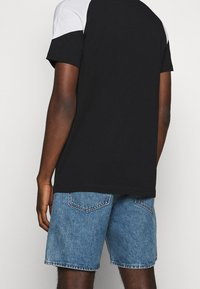 Muž viděný zezadu, který má na sobě černo-bílé tričko s krátkým rukávem a modré džínové kraťasy na jednoduchém světlém pozadí.