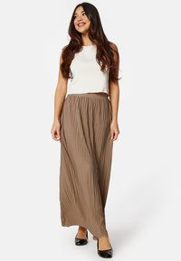 Crème mouwloze top gecombineerd met een lange, geplooide bruine rok met een hoge taille. De rok heeft een zachte textuur en valt tot aan de enkels.