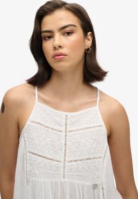 Witte jurk met complexe kantdetails, korte schouderbandjes, een vloeiende rok en een decoratieve zilveren accent aan de onderkant.