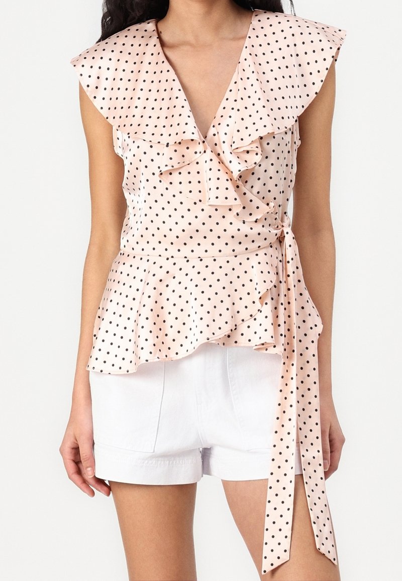 Blouse cache-cœur rose clair sans manches à pois noirs, col et ourlet à volants, nouée à la taille, portée avec un short blanc.