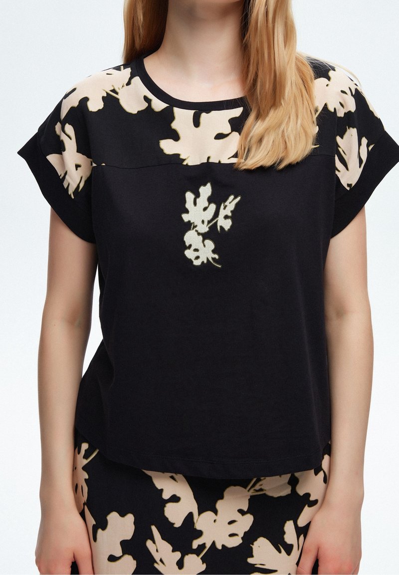 Femme portant un haut noir et une jupe avec des motifs de feuilles beiges, montrant la zone médiane du corps ainsi qu'une partie des bras et du cou.
