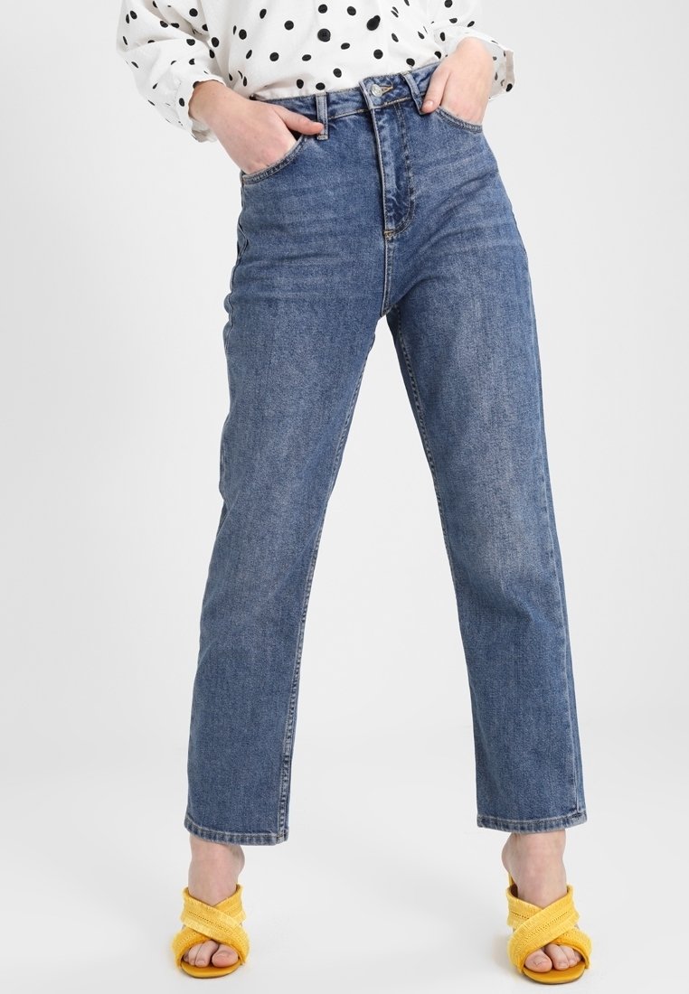 Personne portant un jean droit taille haute bleu avec les mains dans les poches, un chemisier blanc à pois noirs, et des sandales jaunes à bouts ouverts.