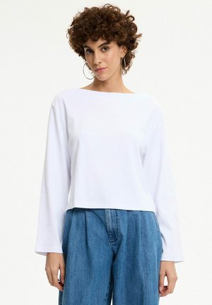 Mujer con cabello rizado que lleva pendientes grandes de aro, una camiseta blanca de manga larga y pantalones vaqueros azules holgados, de pie frente a un fondo blanco liso.